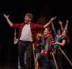 Crane in red checkered work shirt, white t-shirt, black jeans, backward baseball cap holds up his hands as he strides before a rabble of cheering people, all with wooden swords.